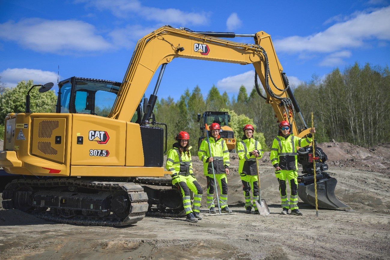 Fyra elever i gula varselkläder står intill en gul grävmaskin på vad som ser ut som ett övningsområde med skog i bakrunden och klarblå himmel med lätta vita moln.