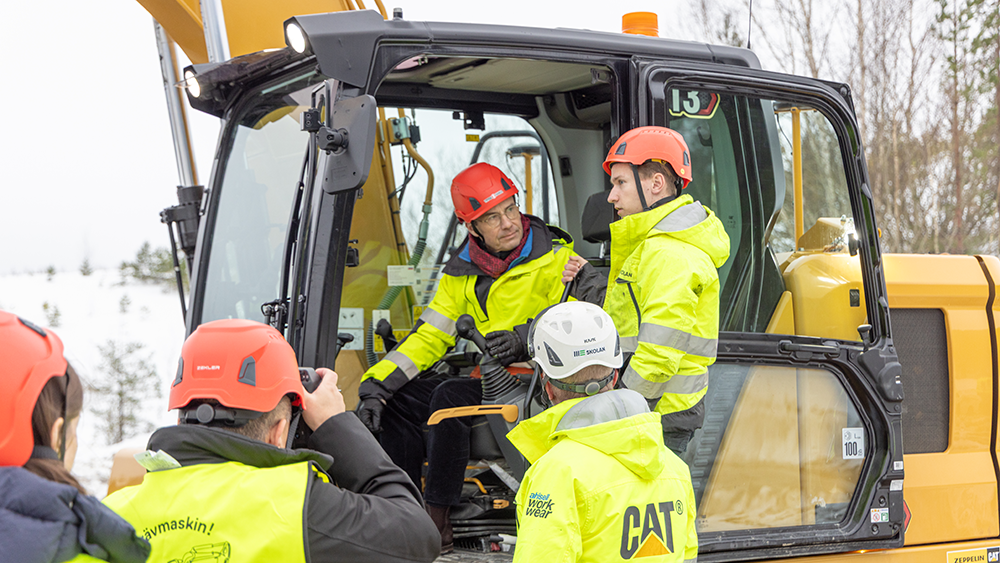 Satsminister Ulf Kristersson iklädd varselkläder får instruktioner i grävmaskinen av elev på ME-skolan i Bålsta