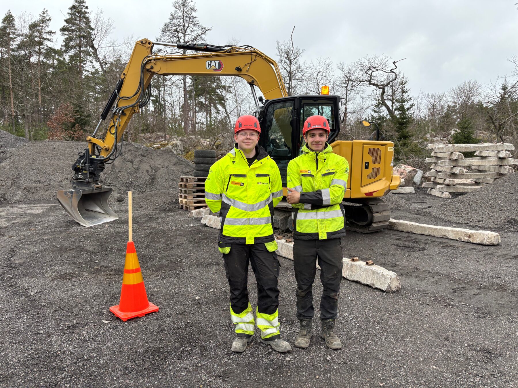 Rasmus och Jonny i varselkläder och hjälmar framför en gul CAT-grävmaskin